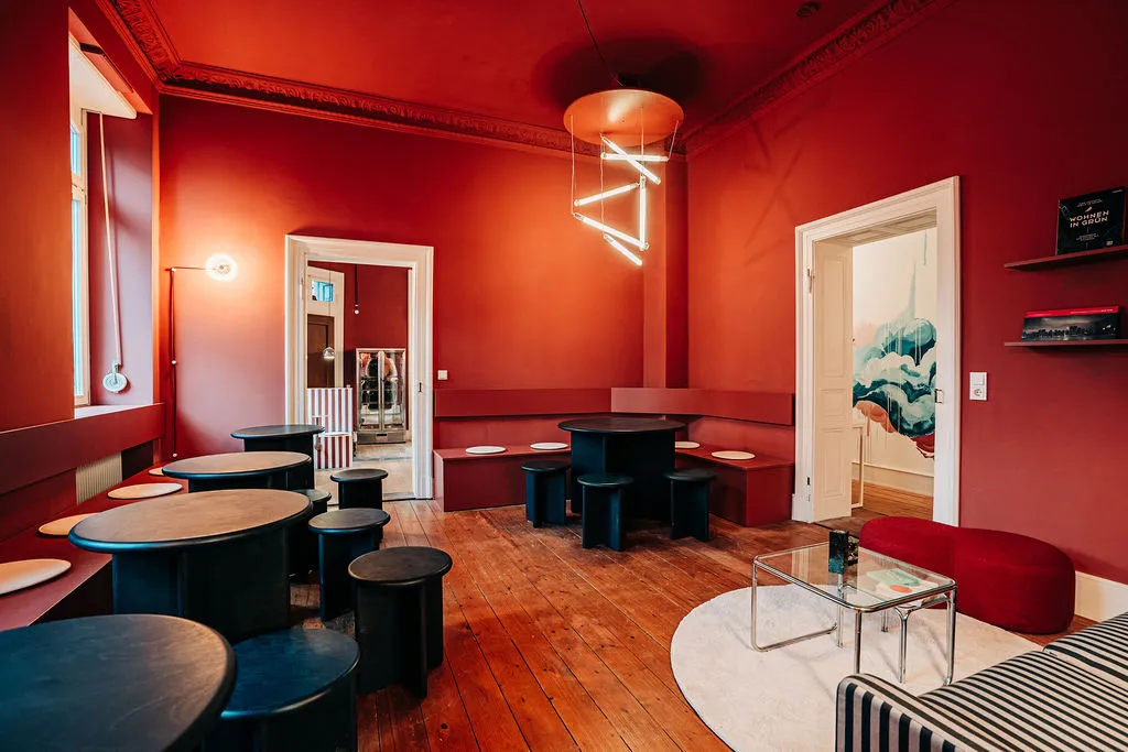 Café-Raum mit roten Wänden, dunkelblauen Tischen, Neonleuchte und Blick ins Wohnzimmer
